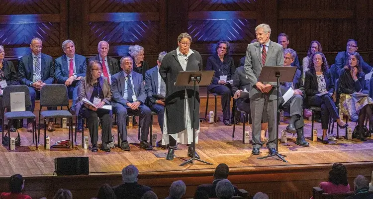 Several individuals are seated on stage behind two people standing at podiums. At the podium on the left is Anna Deavere Smith, dressed in a long black sweater over a white dress and reading from materials on the podium.  To her right, standing at the other podium is James Fallows, who is wearing a gray business suit and is looking at Smith.