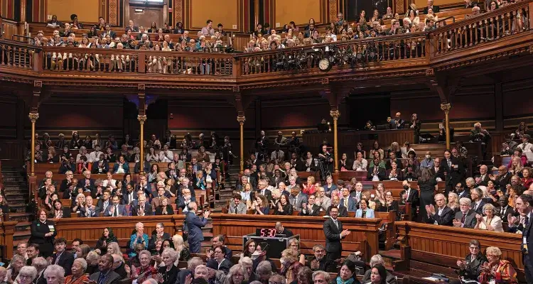 An image of the inside of Sanders Theatre at Harvard University, with people seated throughout the theatre smiling and facing the stage.