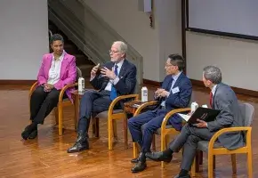 Four individuals are seated next to each other and facing an audience that is not shown.  The second person from the left is speaking and has his hands raised to emphasis a point he is making.  The other three individuals are turned and facing the person who is speaking.