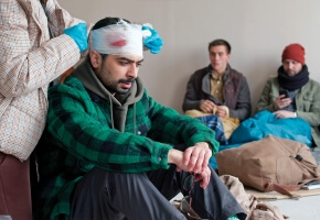 An adult sits on the floor beside a backpack. They have pale skin, a thick dark mustache, and short black hair. A person stands behind them and bandages the top of their head. Red can be seen through the bandages. Two other people wait in the background.